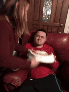 a man in a red shirt is laying on a couch while a woman holds a plate of food over his head