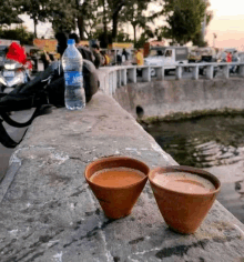 two cups of tea are sitting on a concrete wall next to a bottle of water .