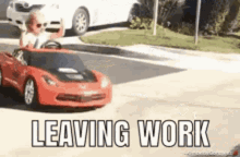 a little boy is driving a red toy car on a street with the words leaving work below him .