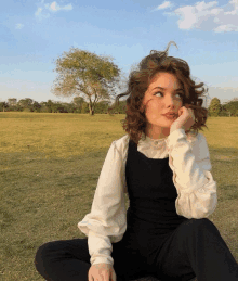 a woman in overalls and a white shirt sits in a field