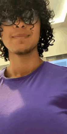 a young man with curly hair and glasses is wearing a purple shirt and smiling .