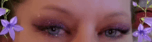 a close up of a woman 's eyes with purple eyeshadow and purple flowers in the background