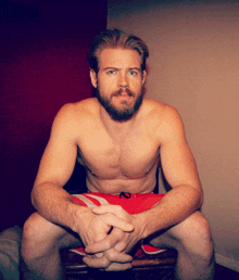 a shirtless man with a beard sits on a stool with his hands folded