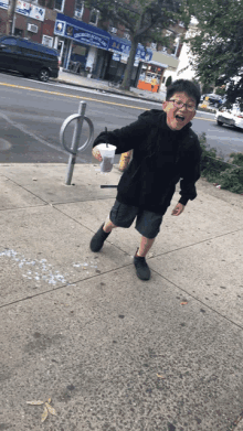 a young boy is standing on a sidewalk holding a cup with a drink in it