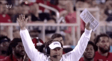 a football coach is wearing headphones and holding a piece of paper in his hand while watching a game .