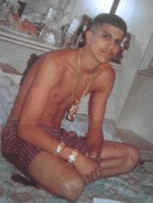 a shirtless man is sitting on a bed wearing shorts and a gold chain .