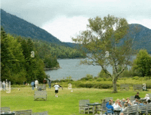 a group of people sit at tables in a grassy area with a lake in the background