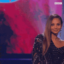 a woman in a black sequined dress is smiling in front of a pink background with bbc written on it