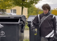 a man wearing a black shirt with the word sall on it is standing next to two trash cans .