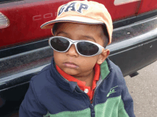 a young boy wearing sunglasses and a gap hat stands in front of a red car