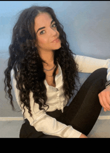 a woman with curly hair wearing a white shirt and black pants sits on the floor