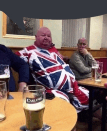 a man in a british flag outfit sits at a table next to a carling glass of beer