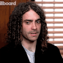 a man with long curly hair and a beard is standing in front of a window with the word billboard behind him