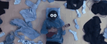 a person in a blue hoodie with a mask on laying on the floor surrounded by clothes