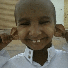 a young boy with a shaved head and big ears is smiling and covering his ears with his hands .