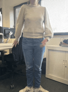 a woman wearing a sweater and jeans stands in front of a desk