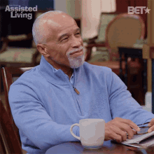 a man is sitting at a table with a cup of coffee and a newspaper with assisted living written on the screen behind him