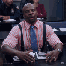 a man in a pink and blue striped shirt and tie sits at a desk with his hands folded in front of his face