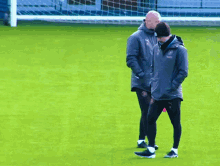 two men walking on a soccer field with one wearing a jacket that says ' manchester city ' on it