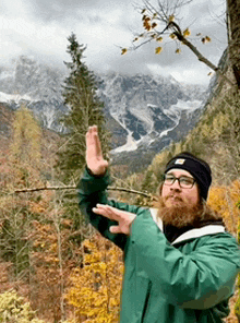 a man with a beard and glasses is standing in front of a mountain with his hands in the air