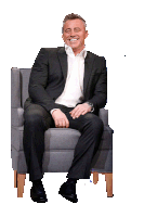 a man in a suit and white shirt is sitting in a chair and smiling