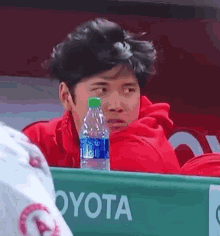 a man in a red jacket is sitting in the dugout with a bottle of water next to him .