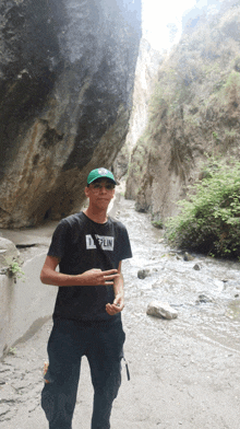 a young man wearing a black shirt that says merlin stands in front of a river