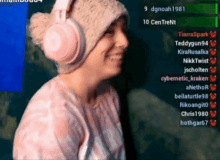 a woman wearing pink headphones and a pink beanie is smiling in front of a screen that says dgnoah1981