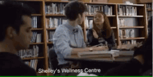 a group of people are sitting at a table in a library with the words shelley 's wellness centre written on the screen .