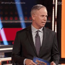 a man in a suit and tie stands in front of a microphone holding a sign that says family feud canada