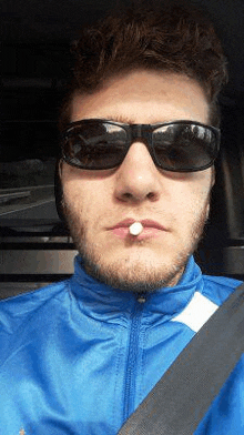 a man wearing sunglasses and a blue jacket is sitting in a car with a pill in his mouth .