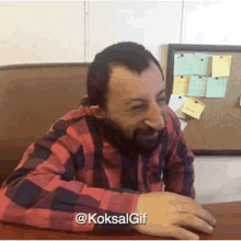 a man with a beard is sitting at a table in front of a bulletin board with sticky notes on it .