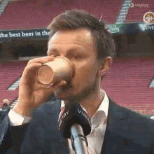 a man in a suit is drinking from a paper cup in front of a sign that says " the best beer in the "