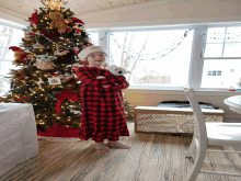 a little girl in a red and black plaid dress holds a stuffed animal in front of a christmas tree