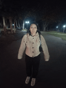 a woman wearing a white jacket and black pants stands in a dark park at night