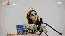 a woman wearing sunglasses and a headband with googly eyes sits in front of a microphone holding a bag of food