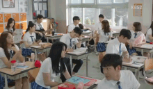 a group of students are sitting at desks in a classroom with kbs2 written on the wall behind them .