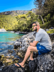 a man wearing sunglasses sits on a rock near a body of water