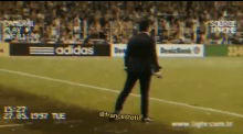 a man in a suit is walking on a soccer field with an adidas sign in the background .