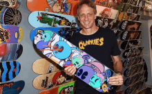 a man wearing a shirt that says quackies is holding a skateboard in front of a wall of skateboards