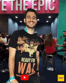 a man wearing a shirt that says " ready for war " stands in front of a sign that says " be the epic "