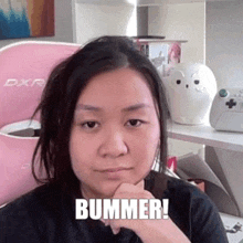 a woman is sitting in front of a pink chair with her hand on her chin and the words bummer written on her face .