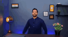 a man in a blue shirt is standing in front of a desk with a laptop and a potted plant .