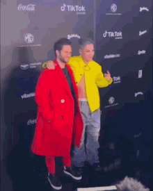 two men are posing for a picture in front of a wall that has coca cola and tik tok written on it