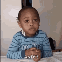 a young boy in a blue striped shirt is sitting at a table with his hands folded and making a funny face .