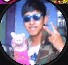 a man wearing sunglasses and a camouflage jacket is holding a stuffed animal and giving a peace sign .