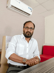 a man wearing glasses and a watch sits at a table in front of a wall mounted air conditioner