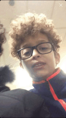 a young boy with curly hair wearing glasses and a blue jacket with an orange collar