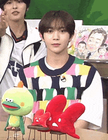 a young man sitting at a table with stuffed animals and a drawing of a man on the wall behind him