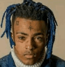 a man with blue dreadlocks and a tattoo on his face is wearing a denim jacket and a sweater .